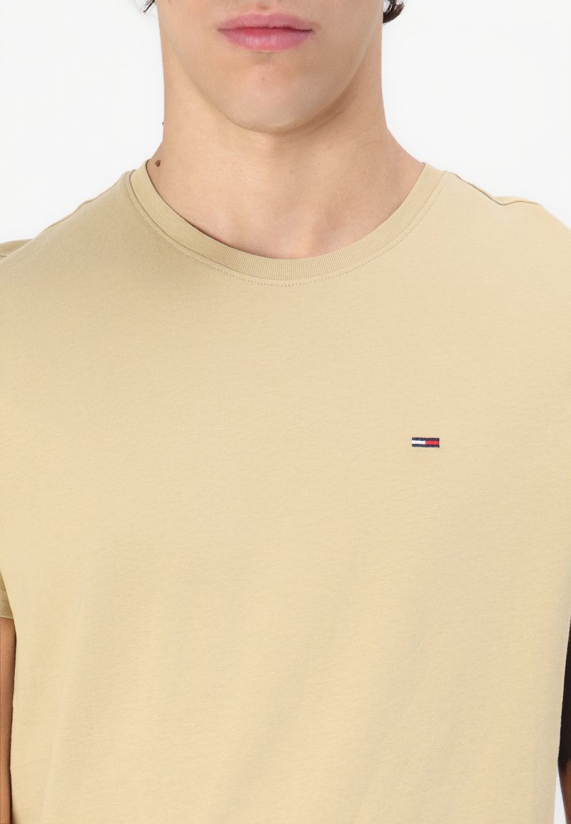 Camiseta de algodón beige con escote redondo y mangas cortas, que presenta un pequeño logotipo bordado en rojo, blanco y azul en el pecho.