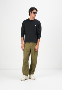 Uomo che indossa una maglietta nera a maniche lunghe, pantaloni cargo verde oliva, sneakers beige e occhiali da sole neri, in piedi contro uno sfondo bianco semplice.