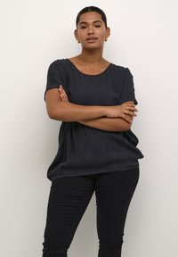 Top nero a maniche corte con scollatura rotonda e vestibilità comoda, realizzato in tessuto testurizzato; abbinato a pantaloni skinny neri; sfondo neutro.