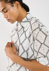 Kurzarmhemd aus weißer Spitze mit schwarzen Rautenmustern. Verfügt über einen klassischen Kragen und eine lockere Passform. Strukturierter Stoff mit filigranen Details.