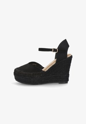 Zapato de esparto negro con cuña, con suela de yute trenzado, punta redondeada y correa en el tobillo. Parte superior de tela suave y detalles texturizados en el talón.