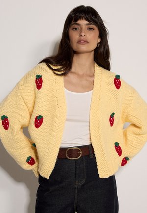 Mujer con un cárdigan amarillo de punto con patrones de fresas rojas sobre una camiseta blanca, combinado con jeans oscuros y un cinturón marrón.