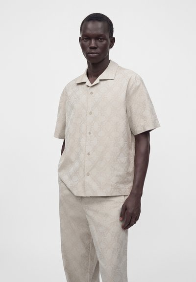 Homme portant une chemise beige à manches courtes boutonnée et un pantalon assorti à motif, debout devant un fond blanc uni.