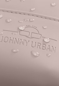 A világos rózsaszín vízálló anyag, amelyen kiemelkedő "JOHNNY URBAN" logó látható, vízcseppekkel a felületén, amelyek a vízállósági tulajdonságait jelzik.