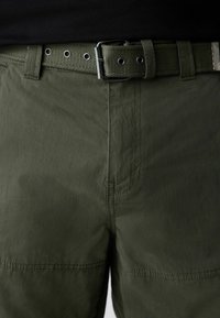 Close-up of dark green pants with matching fabric belt featuring metal buckle and eyelets, worn with a black top.