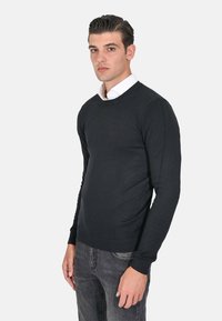 Maglione nero a maniche lunghe con scollo rotondo, realizzato in un morbido tessuto a maglia. Presenta polsini e orlo a coste, indossato sopra una camicia bianca con colletto.