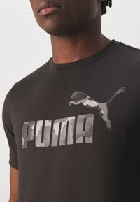 Černé tričko s krátkým rukávem, které na přední straně obsahuje velké šedé kamuflážové logo "PUMA" a doprovodnou grafiku pumu skákající v podobných odstínech.