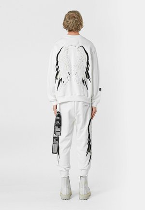 Sudadera de algodón blanca con bordado de alas en negro y plata en la parte posterior, combinada con pantalones de chándal blancos que presentan etiquetas impresas y rayas laterales.