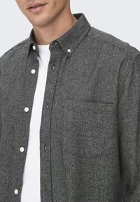 Chemise grise à manches longues en tissu texturé, dotée d'un col, de boutons blancs et d'une poche poitrine unique sur le côté gauche.