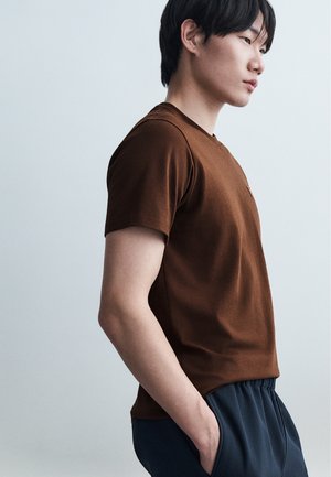 Νεαρός άνδρας σε προφίλ, φοράει καφέ μπλουζάκι με κοντά μανίκια και σκούρο παντελόνι, με το δεξί του χέρι στην τσέπη.