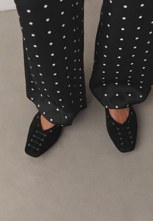 Čierne nohavice s bodkami spárované s čiernymi špicatými topánkami so elastickými viazacimi detailmi na šedej podlahe.