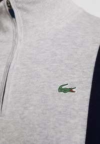 Pull gris clair en maille avec une fermeture éclair et une bande bleu marine, arborant un petit logo de crocodile vert sur la poitrine.