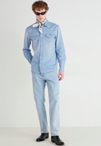 Camisa de mezclilla azul claro con botones a presión, dos bolsillos en el pecho, combinada con pantalones vaqueros azul claro. Gafas de sol negras y una bufanda con estampado añaden detalle.