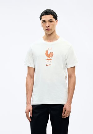 Vyras dėvi baltą marškinėlį su oranžiniu gaidžio emblematu, dviem žvaigždutėmis, „FFF“ raidėmis ir Nike logotipu, derinamus su juodomis kelnėmis.