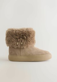 Botine din suede bej, cu manșetă din blană sintetică, legate cu o funda și accentuate de două clopoței de culoare aurie. Talpă din cauciuc plat pentru confort.