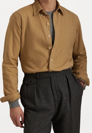 Mand iført en kamelfarvet skjorte med knapper over en grå sweater, kombineret med højtaljede, mørkegrå plisserede bukser, hænder delvist i lommerne.