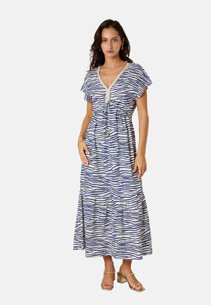 Femme portant une robe longue à motif zèbre bleu et blanc, avec un décolleté en V et des manches courtes, assortie à des sandales à talons blocs dorés.