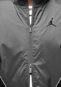 Grå vindjacka med framdragkedja, med en subtil Jordan-logotyp på bröstet, tillverkad av ett mjukt, lätt material.
