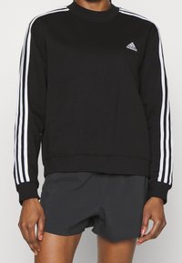 Svart sweatshirt med vit tre-stripsdesign på ärmarna, rundad hals, ribbade muddarna och liten Adidas-logotyp på bröstet.