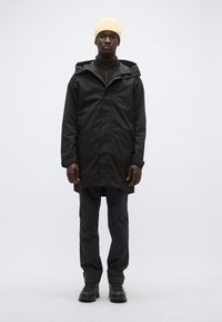 Μαύρο αδιάβροχο παρκά με κουκούλα, μπροστινό καβάλο και πλαϊνές τσέπες. Συνδυάζεται με μαύρο παντελόνι και μαύρα αθλητικά παπούτσια. Κίτρινο πλεκτό σκουφάκι.