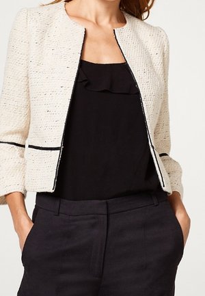 Kobieta w kremowej, teksturowanej, krótkiej kurtce z czarną lamówką, na czarnej bluzce z falbanką przy dekolcie i czarnych spodniach z wysokim stanem, z rękami w kieszeniach.
