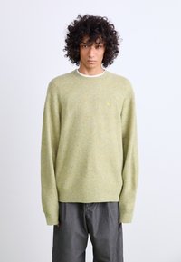 Maglione a maglia verde chiaro con una texture morbida, scollatura e polsini a costine, caratterizzato da un piccolo logo sul petto, abbinato a pantaloni grigi.