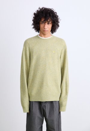 Suéter de punto verde claro con una textura suave, cuello y puños acanalados, que presenta un pequeño logo en el pecho, combinado con pantalones grises.