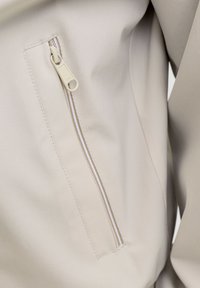 Ανοιχτό μπεζ μπουφάν με πλαϊνή τσέπη με φερμουάρ σε λευκό χρώμα και υφασμάτινη υφή, που παρουσιάζει καθαρές γραμμές και μοντέρνο σχεδιασμό.