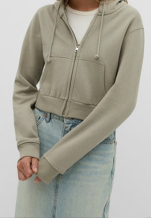 Femme portant un sweat à capuche court beige clair zippé sur un haut blanc et une jupe en jean bleu clair, les mains jointes devant.
