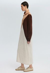 Robe longue à blocs de couleurs avec un décolleté drapé. Les matériaux incluent du tissu marron, beige et crème au toucher doux. Portée avec des chaussures beiges.