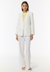 Weißer Leinenblazer mit einem einzigen Knopf, zwei Fronttaschen, kombiniert mit weit geschnittenen weißen Leinenhosen und einem hellgelben Oberteil. Schlichte Schuhe.