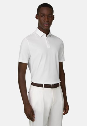 Homme portant un polo blanc à manches courtes rentré dans un pantalon blanc avec une ceinture marron foncé, debout devant un fond uni.