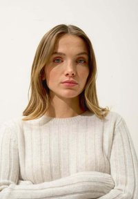 Pull en tricot côtelé beige clair, avec un col rond et des manches longues, présentant une texture douce et une coupe décontractée. Le modèle pose les bras croisés.