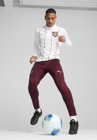 Balta ilga rankovė viršutinė su raudonomis juostelėmis ir logotipu; bordo liesi kelnės su logotipu; juodi futbolo bateliai; mėlynas ir baltas futbolo kamuolys.