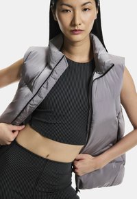 Grå polstret vest med høj krave, lynlåslukning og lomme, båret over en sort stribet cropped top og matchende bund. Glat, skinnende tekstur.