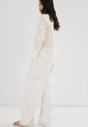 Femme aux longs cheveux foncés portant un chemisier blanc transparent à motifs et un pantalon blanc à jambes larges, debout de profil devant un fond uni.