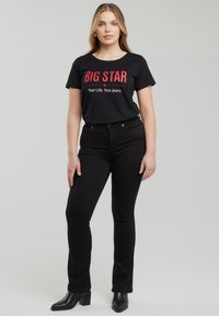 Nő áll, fekete "BIG STAR Your Life. Your Jeans." feliratú pólót, fekete szűk farmernadrágot és fekete magassarkú csizmát visel világosszürke háttér előtt.