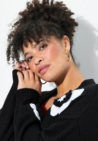 Schwarzer Strickpullover mit einer weißen Blumenapplikation auf der Schulter, gerippten Bündchen und einer glatten Textur. Model posiert mit den Händen im Gesicht.