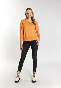 Oranger Pullover mit langen Ärmeln, kombiniert mit schwarzen, engen Jeans und cremefarbenen Stiefeletten mit schwarzen Sohlen und Schnürsenkeln. Das Model steht selbstbewusst.