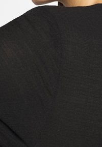 Prenda de tela negra texturizada con un diseño holgado, que muestra líneas suaves y drapeadas, y un ligero brillo en la superficie.
