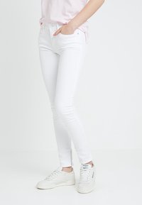 Persona vestida con jeans blancos ajustados, camisa rosa claro y zapatillas Reebok blancas, de pie con una mano en el bolsillo sobre un fondo blanco.