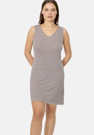 Mujer con vestido sin mangas hasta la rodilla con patrón de zigzag rojo, verde y blanco, de pie con las manos detrás de la espalda sobre un fondo blanco.
