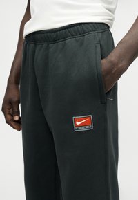 Κοντινό πλάνο ενός ατόμου που φοράει μαύρο σορτς Nike με ελαστική μέση και πλαϊνές τσέπες, ένα χέρι στην τ σέπη, λευκό πουκάμισο μερικώς ορατό.