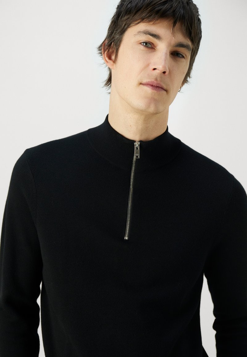 Young man wearing a black long-sleeve sweater with a silver front zipper, looking slightly to the side against a plain white background.