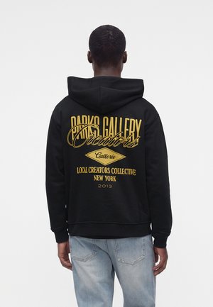 Person, die einen schwarzen Hoodie mit gelbem Text ""Paris Gallery Creators Gallerie Local Creators Collective New York 2013"" auf dem Rücken trägt.
