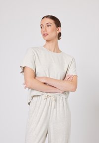 Žena s vlasy staženými do drdolu, oblečená v lehkém béžovém tričku s krátkými rukávy a kalhotách se šňůrkou v pase, s překříženýma rukama, dívající se vlevo na jednobarevném pozadí.