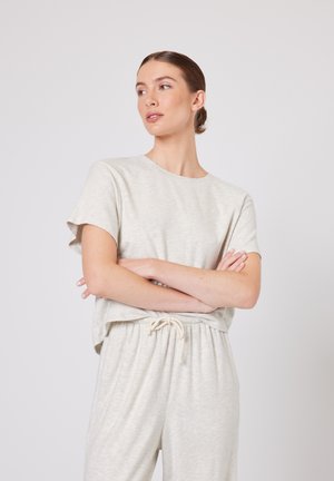 Žena s vlasy staženými do drdolu, oblečená v lehkém béžovém tričku s krátkými rukávy a kalhotách se šňůrkou v pase, s překříženýma rukama, dívající se vlevo na jednobarevném pozadí.