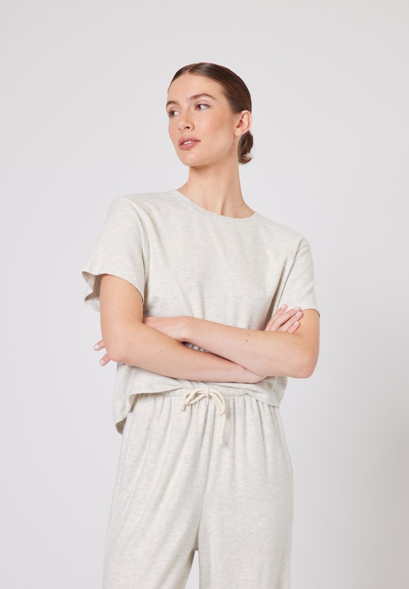 Žena s vlasy staženými do drdolu, oblečená v lehkém béžovém tričku s krátkými rukávy a kalhotách se šňůrkou v pase, s překříženýma rukama, dívající se vlevo na jednobarevném pozadí.