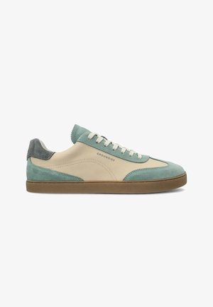 Low-top sneaker met crème leer en teal suède overlays, witte veters, bruine zool en "GROUNDIES" branding aan de zijkant.