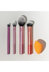 Cinq outils de maquillage Real Techniques, quatre pinceaux avec des manches métalliques roses et orange, et une éponge de maquillage orange sur une surface blanche.